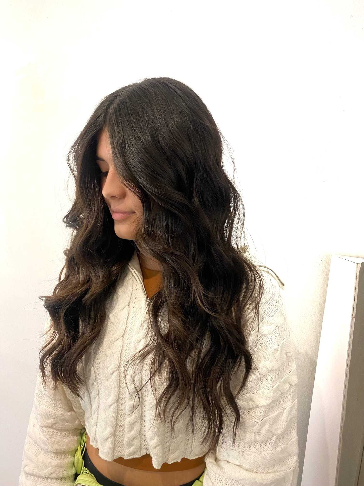 Young woman with long, wavy dark hair, wearing a white textured jacket, posing sideways.